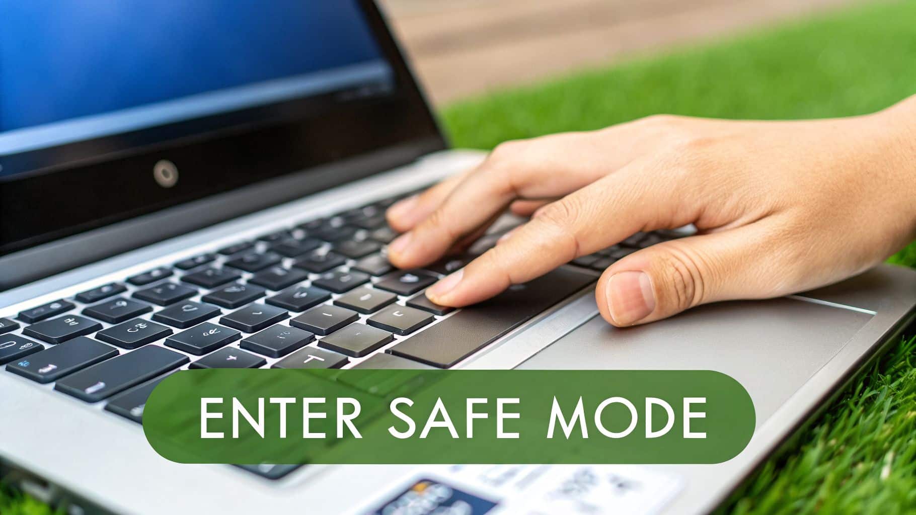 A close-up of a hand typing on a laptop keyboard on green grass, with 'ENTER SAFE MODE' overlay.