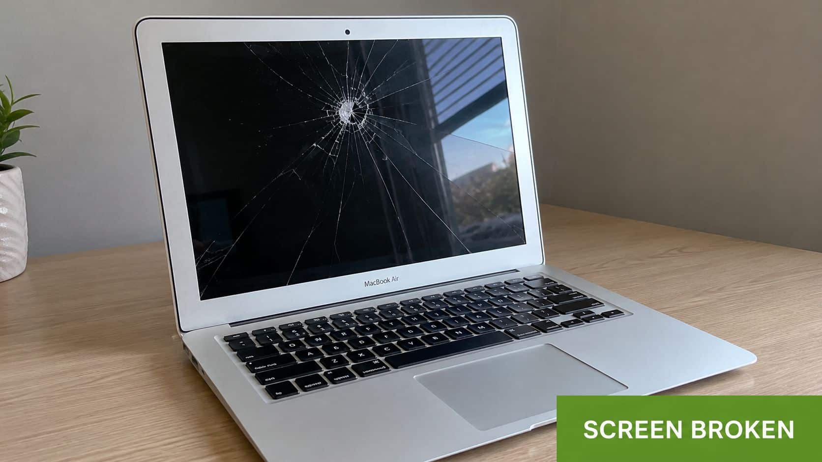 A silver MacBook Air laptop sitting on a wooden desk with a badly cracked and broken screen.