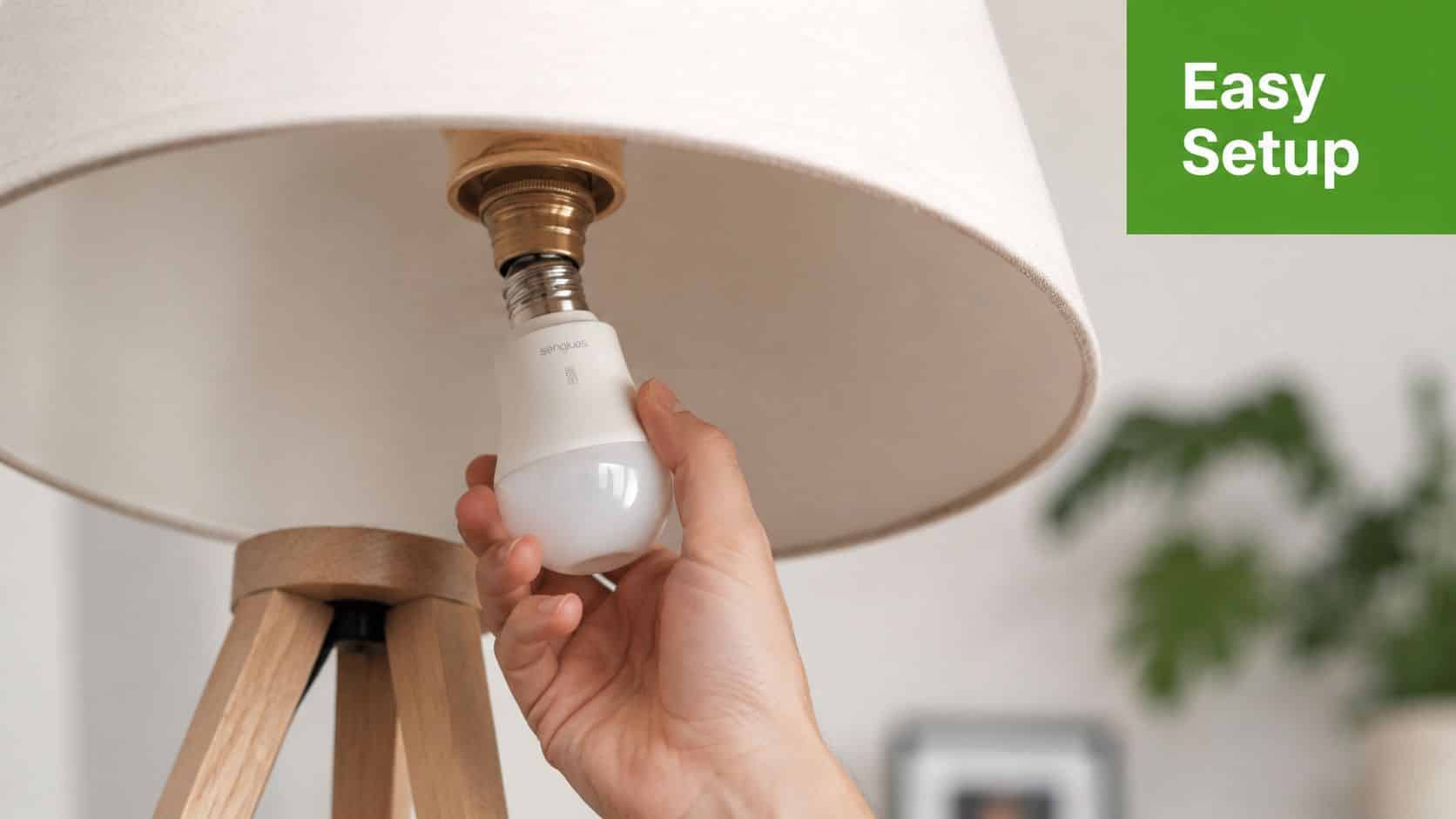 A hand installing a white smart light bulb into the socket of a wooden floor lamp.